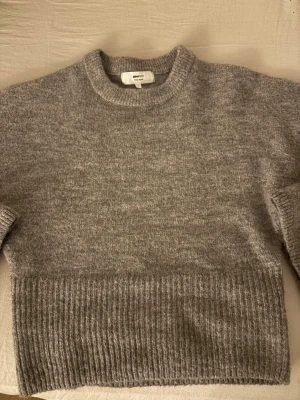 Grå stickad tröja från Gina Tricot - Mysig grå stickad figur formad tröja från Gina Tricot i ullblandning. Tröjan har rund halsringning, långa ärmar och bred ribbstickad kant nedtill. Perfekt för kyliga dagar och enkel att matcha med olika outfits.