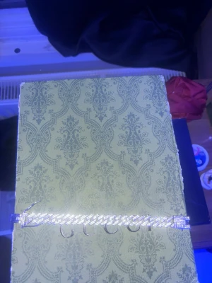 Silverfärgat armband  - Säljer ett stilrent silverfärgat armband med bred kedja och glittrande detaljer. Armbandet har en klassisk kubansk länkdesign och ett lås dekorerat med små stenar för extra bling. Perfekt accessoar för dig som gillar exklusiv och trendig stil.