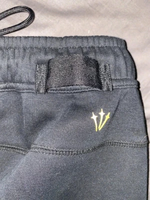 Svarta mjukisbyxor från Nike NOCTA - Svarta mjukisbyxor från Nike NOCTA med elastisk midja och breda hällor. Byxorna har NOCTA-logga med tre stjärnor i gult framtill. Tillverkade i mjukt material som känns skönt mot huden. Perfekta för en avslappnad och trendig stil.