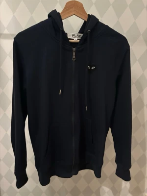 Mörkblå hoodie från Comme des Garçons - Snygg mörkblå hoodie med dragkedja från Comme des Garçons PLAY. Har huva med dragsko, fickor fram och det ikoniska hjärtat med ögon på ryggen samt en liten logga på bröstet. Perfekt för dig som gillar streetwear och vill sticka ut lite extra.