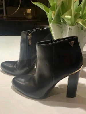 Svarta stövletter från Guess - Snygga svarta stövletter från Guess  med gulddetaljer vid klacken och dragkedja på sidan. Klassisk spetsig tå och hög blockklack ger en stilren och trendig look. Perfekta för att lyfta vilken outfit som helst.