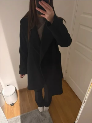 Lång svart kappa - Stilren svart kappa med bred krage och rak passform. Kappan har långa ärmar och går ner till knäna. Perfekt för dig som vill ha en klassisk och tidlös look. Passar till många olika outfits och håller dig varm under kyliga dagar.