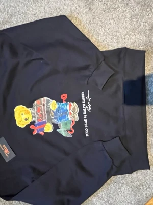 Svart Polo Bear hoodie från Ralph Lauren - Snygg svart hoodie från Ralph Lauren med den ikoniska Polo Bear-trycket framtill. Björnen har på sig en stickad tröja och jeans, och detaljer i rött och blått gör hoodien extra unik. Mjuk bomullskvalitet och klassisk passform med känguruficka.