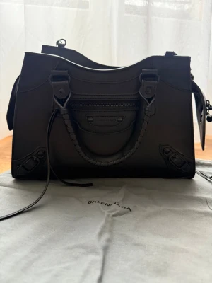 Svart Balenciaga handväska  - Säljer en svart handväska från Balenciaga i slätt läder med klassiska detaljer och flätat handtag. Väskan har både axelrem och handtag, samt en liten ficka med dragkedja framtill. Perfekt för dig som gillar stilrena och exklusiva accessoarer.