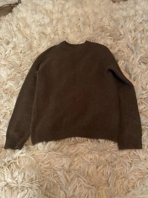 Mörkbrun stickad tröja - En enkel och stilren mörkbrun stickad tröja med rund halsringning och ribbade muddar vid ärmslut och nederkant. Perfekt för lager på lager och passar till det mesta. Materialet är i lambswool blend. Lite nopprig men inget som är tydligt 
