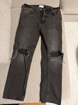 Svarta slitna jeans med hål på knäna - Säljer ett par svarta jeans med raka ben och stora slitningar över båda knäna. Jeansen har fem fickor och klassisk knappgylf. Materialet är denim i bomull och färgen är tvättad svart för en cool, avslappnad look.
