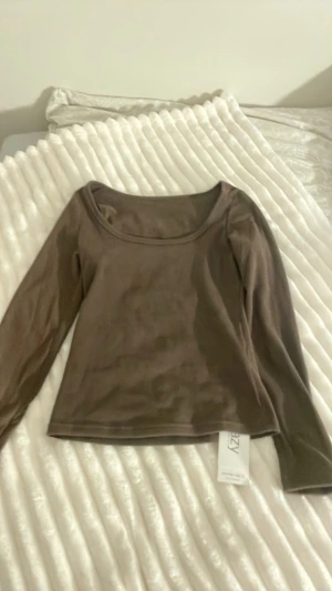 Brun långärmad topp från NA-KD - Mörkbrun långärmad topp med rundad, bred urringning. Tillverkad i mjuk och stretchig polyester som ger en slim och bekväm passform. Perfekt att styla med jeans eller kjol för en clean look.