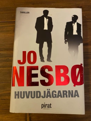 Huvudjägarna av Jo Nesbø - Spännande thrillerroman av Jo Nesbø med titeln Huvudjägarna. Omslaget är vitt med stora röda och grå bokstäver samt silhuetter av två män i kostym. Boken är inbunden och texten är på svenska. Perfekt för dig som gillar nervkittlande berättelser. Inbunden.