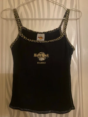Hard Rock Café leopardlinne svart - Svart linne från Hard Rock Café Dubai med leopardmönstrade kanter och axelband. Linnet har broderad logga framtill och är gjort i 100% bomull. Insydd så snarare storlek XS-S