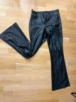 Svarta utsvängda byxor i fuskskinn - Snygga svarta byxor i fuskskinn med utsvängda ben och hög midja. Byxorna har bälteshällor och dragkedja fram. Perfekta för dig som vill ha en edgy och trendig look.