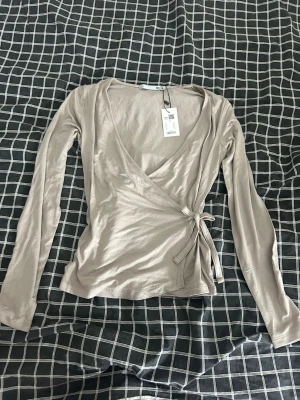 Beige omlottopp med knyt från Bodil - Snygg beige topp med långa ärmar och knyt i sidan. Toppen har en v-ringad design och är gjord i ett mjukt och skönt material som känns skönt mot huden. 