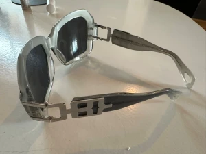 Silvriga fyrkantiga solglasögon - Unika solglasögon ser ut att vara från Jean Paul Gaultier med silvrig metallbåge och mörka, fyrkantiga linser. Sidorna har en cool, industriell design med utskurna detaljer. Perfekt för dig som vill sticka ut med en futuristisk accessoar.