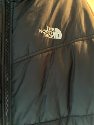Svart pufferjacka från The North Face - Säljer en svart pufferjacka från The North Face med dragkedja framtill och hög krage. Jackan har diskret logga på bröstet och är vadderad för extra värme. Perfekt för kalla vinterdagar och har en klassisk, stilren look.
