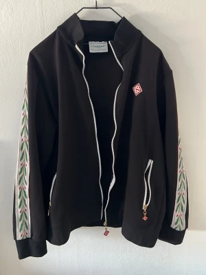 Svart tracksuit Casablanca passar mer storlek M - Svart hoodie från Casablanca med vita dragkedjor och broderade detaljer i grönt och rött längs ärmarna. Märkets logga sitter på bröstet. Hoodien har en modern passform och är tillverkad i mjukt material, perfekt för en avslappnad och trendig look.