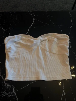 Vit ribbad bandeau tubtopp - Säljer en snygg vit bandeau tubtopp med ribbad struktur och twistad detalj framtill. Perfekt att styla till jeans eller kjol för en trendig look. Toppen är axelbandslös och har en croppad passform.