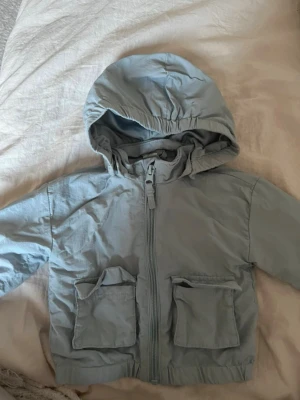 Ljusblå vindjacka med huva - Ljusblå vindjacka i tunt, lätt material med dragkedja framtill och två stora fickor med lock. Jackan har elastisk nederkant och muddar vid ärmsluten samt en rymlig huva. Perfekt för blåsiga dagar och enkel att packa ner i väskan.