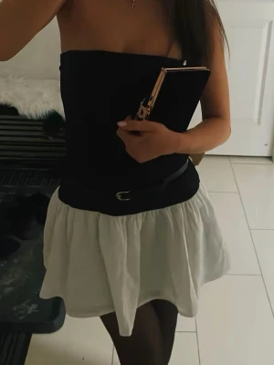 Svart och vit axelbandslös klänning - Trendig axelbandslös klänning med svart överdel och vit, fluffig kjol. Klänningen har ett bälte i midjan som markerar siluetten och ger en snygg kontrast mellan över- och underdel. Perfekt för dig som gillar en stilren och modern look.aldrig använd bara prövad 