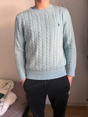 Ljusblå kabelstickad tröja Polo Ralph Lauren - Kabelstickad ljusblå tröja från Polo Ralph Lauren med rund halsringning och diskret logga broderad på bröstet. Tröjan har lång ärm och ribbade muddar vid ärmslut och nederkant. Perfekt för en stilren och klassisk look.