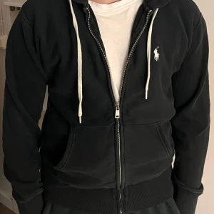 Svart hoodie från Polo Ralph Lauren  - Svart hoodie från Polo Ralph Lauren med vit broderad logga på bröstet. Tröjan har huva med vita snören, dragkedja framtill och två stora fickor. Mjuk bomullskvalitet  Perfekt för chill dagar. Står S men passar som xxs