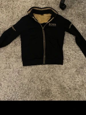 Svart hoodie från BOSS med gulddetaljer - Snygg svart hoodie från BOSS med guldfärgade detaljer och dragkedja framtill. Loggan är broderad på bröstet och insidan av huvan är beige. Stilsäkra detaljer längs ärmar och huva ger en exklusiv känsla. Perfekt för dig som vill ha en clean men lyxig look.