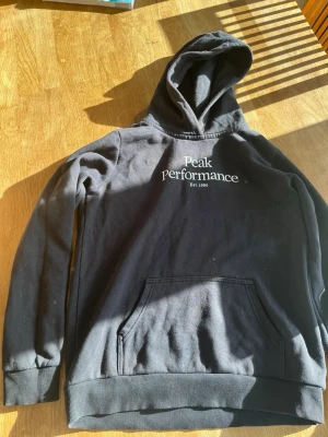 Svart hoodie Peak Performance - Snygg svart hoodie från Peak Performance med vit logga och text på bröstet. Klassisk känguruficka framtill och ribbade muddar vid ärmar och nederkant. Tillverkad i mjukt bomullsmaterial och har en stor huva för extra komfort. Jag har använt den typ 2 gånger och man kan inte se att den är använd. Ny kostar 800 