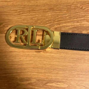 Svart och brunt bälte med guldspänne - Snyggt bälte från Ralph Lauren med ett stort guldspänne som har RL-loggan. Bältet är vändbart med en sida i svart och den andra i brunt, vilket gör det mångsidigt för olika outfits.