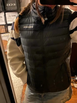 Svart dunväst från Stormberg - Snygg svart dunväst från Stormberg med hög krage och dragkedja framtill. Västen har två fickor med dragkedja och diskret logga på bröstet. Perfekt att slänga över en hoodie eller tröja för extra värme. Lätt och smidig design som funkar året runt.