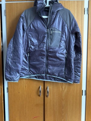 Grå pufferjacka från Peak Performance - Snygg grå pufferjacka från Peak Performance med huva och dragkedja framtill. Jackan har en bröstficka med dragkedja och logga, samt två sidofickor. Materialet är glansigt och lätt vadderat, perfekt för kalla vinterdagar.