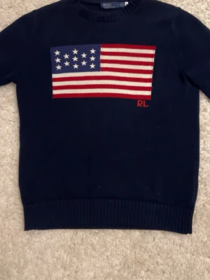 stickad tröja Polo Ralph Lauren - Mörkblå stickad tröja från Polo Ralph Lauren med amerikansk flagga i rött, vitt och blått på bröstet. RL-broderi i rött nedtill på flaggan. Rund halsringning och ribbade muddar vid ärmslut och nederkant. Perfekt för en casual look.
