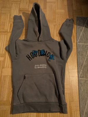 Grå hoodie från Hoodrich med tryck - Säljer en grå hoodie från Hoodrich med stor huva och magficka. Framsidan har Hoodrich-logga i svart och blått samt texten 'From Nothing To Something'. Baksidan har stort tryck med texten 'product of the streets'. Perfekt streetwear-plagg med skön passform.