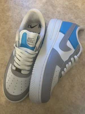 Nike Air Force 1 grå/blå sneakers - Nike Air Force 1 sneakers i vitt, grått och blått. Klassisk låg modell med perforerad tå, vita skosnören och grå swoosh. Ovandelen är i läder och detaljerna i blått ger en fräsch kontrast. Perfekt för dig som gillar stilrena och sportiga sneakers. Bara att höra av sig om ni är intresserade😊