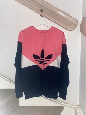 Adidas sweatshirt rosa och svart - Säljer en snygg sweatshirt från Adidas med färgblock i rosa, svart och vitt. Tröjan har det klassiska Trefoil-loggan framtill, rund halsringning och långa ärmar. Perfekt för dig som gillar streetwear och sportig stil.