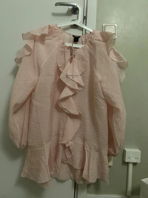  Ny Ljusrosa volangblus från Lindex - Supersöt ljusrosa blus från Lindex med volanger längs fram och vid ärmslut. Blusen har långa ärmar, rund halsringning med knytband och är gjord i ett lätt, krinklat tyg som ger en luftig känsla. Perfekt för dig som gillar romantisk stil.