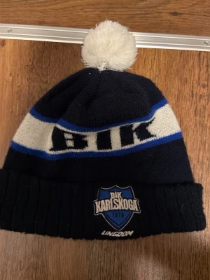 BIK Karlskoga mössa med tofs - Snygg mörkblå och vit BIK Karlskoga mössa med blå detaljer och vit tofs på toppen. Mössan har broderad BIK Karlskoga-logga framtill och bred vikt kant. Perfekt för dig som vill visa ditt stöd för laget under kalla dagar.