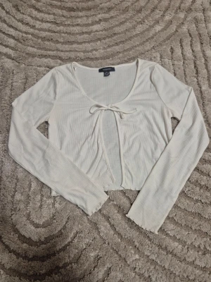 Vit ribbad croppad topp/cardigan från Primark - Trendig vit ribbad topp/cardigan från Primark med lång ärm och croppad passform. Toppen har en öppen front med knytband och rundad urringning. Storlek 34-36. Perfekt att styla med jeans eller kjol för en snygg look. Skriv om prisförslag eller bilder ❤️🫶