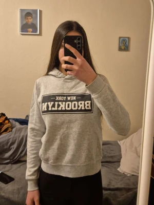 Grå hoodie med Brooklyn-tryck - Snygg grå hoodie med svartvit tryck på bröstet där det står 'New York Brooklyn'. Klassisk passform med huva och ribbade muddar vid ärmslut och midja. Perfekt för en avslappnad streetstyle-look.