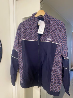 Mönstrad vindjacka från Les Deux - Snygg vindjacka från Les Deux i marinblått med färgglatt geometriskt mönster i rött, blått och vitt på axlar och ärmar. Jackan har hel dragkedja, hög krage och vita detaljer. Perfekt för dig som vill sticka ut med stilren design.