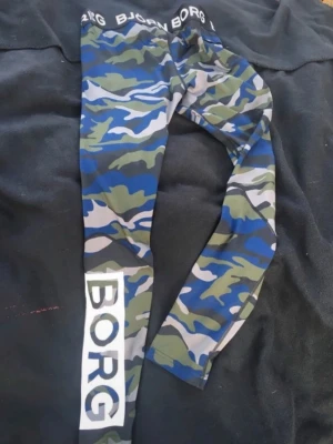 Camo tights från Björn Borg - Snygga träningsbyxor/ tights från Björn Borg med blå, grön och grå kamouflagemönster. Elastisk midja med Björn Borg-logga och stor vit BORG-text på benet. Tillverkade i stretchigt funktionsmaterial som passar perfekt till träning.