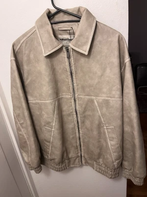 Beige skinnjacka med dragkedja - Stilren beige skinnjacka med klassisk krage och dragkedja framtill. Jackan har två snedställda fickor och elastiska muddar vid ärmslut och nederkant för en ocersized passform. 