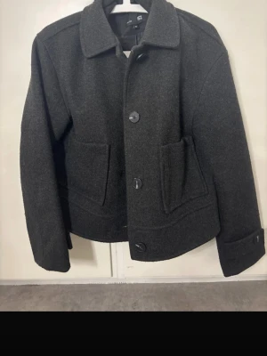 Mörkgrå ullrock från 157, XS - Stilren mörkgrå ullrock från 157 med klassisk krage, stora knappar och två rymliga fickor framtill. Kortare modell som ger en modern look. Perfekt för dig som gillar minimalistisk och tidlös stil. Materialet är tjockt och känns rejält.