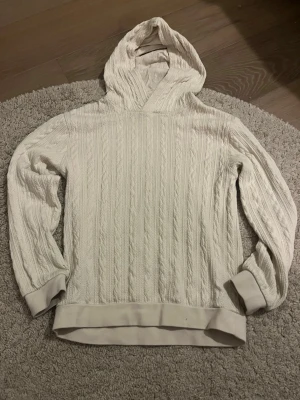 Vit stickad hoodie med kabelmönster - Säljer en krämvit stickad hoodie med klassiskt kabelstickat mönster. Tröjan har huva, långa ärmar och ribbade muddar vid ärmslut och nederkant. Perfekt för dig som gillar en avslappnad men ändå stilren look. Materialet känns mjukt och mysigt.