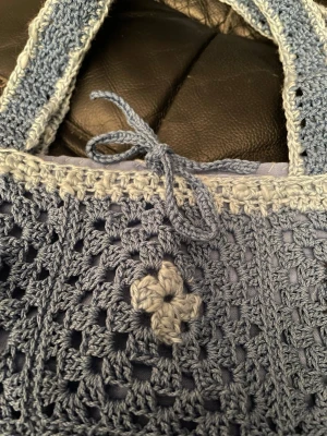 Blå handvirkad shoppingväska - Unik shoppingväska i handvirkad stil med ljusblå och mörkblå garn. Helt ny och unik. Väskan har blommiga detaljer, virkade handtag och en dekorativ knytning upptill. Perfekt för dig som gillar boho och retrovibbar. Materialet är mjukt garn och väskan har en fyrkantig form. Tygfoder inuti så att inte småsaker ska falla ut. 