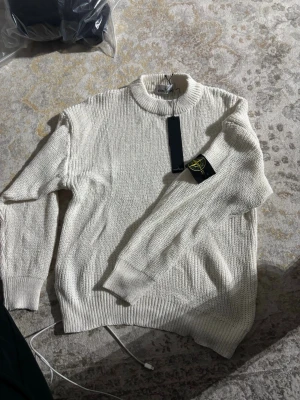 Vit stickad tröja från Stone Island - Snygg vit stickad tröja från Stone Island med ribbad krage och muddar. Tröjan har en klassisk passform och är tillverkad i ett mjukt, grovstickat material. Perfekt för dig som gillar stilrena och trendiga plagg.