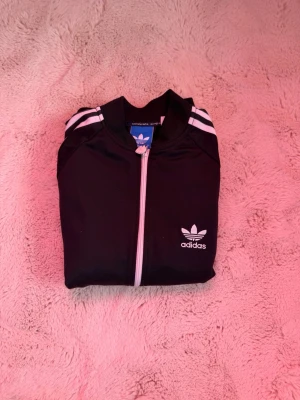 Svart Adidas Originals träningsjacka - Klassisk svart träningsjacka från Adidas Originals med vita ränder längs ärmarna och vit dragkedja. Jackan har ribbade muddar, ståkrage och Adidas logga på bröstet. Perfekt för sport eller streetwear-stil.