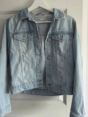 Ljusblå jeansjacka från GinaTricot - Snygg ljusblå jeansjacka från gina med klassisk krage, två bröstfickor med lock och silverfärgade knappar. Jackan har raka ärmar och en tidlös design som passar till det mesta. Perfekt för dig som gillar en avslappnad och trendig stil.