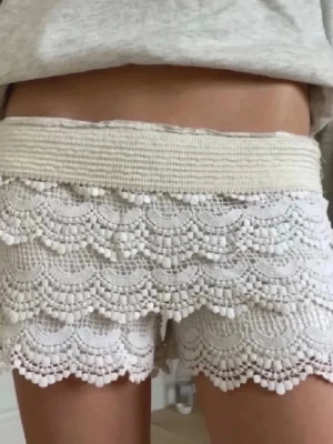 Vita virkade shorts med spetsdetaljer - Säljer ett par vita shorts med bred resår i midjan och flera lager virkad spets. De har en feminin look med vågiga kanter och är perfekta för varma dagar. Supersnygga att matcha med en enkel topp.