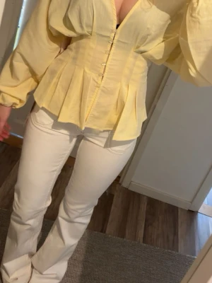 Gul blus med puffärmar från Nelly - Snygg ljusgul blus från Nelly med markerad midja, knappar framtill och voluminösa puffärmar. Blusen har v-ringning och är tillverkad i ett tunt, lätt material som ger en luftig känsla. Perfekt för dig som vill sticka ut med en trendig look. Endast använd 1 gång