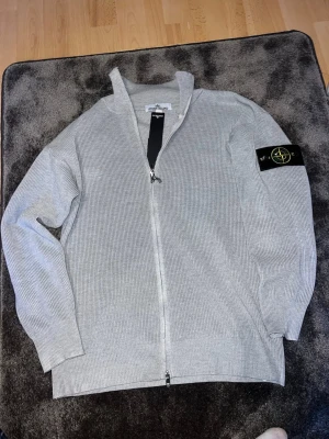 Grå ziptröja från Stone Island - Snygg grå ziptröja från Stone Island med ribbad struktur och ikonisk logotyp-patch på ärmen. Tröjan har hög krage och dragkedja hela vägen. Perfekt för en stilren och avslappnad look.