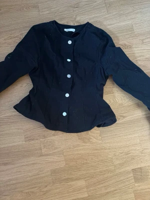 Svart figursydd blus med knappar - Snygg svart blus med figurnära passform och stora vita knappar framtill. Blusen har långa ärmar och rund halsringning. Perfekt till både jeans och kjol för en trendig look.