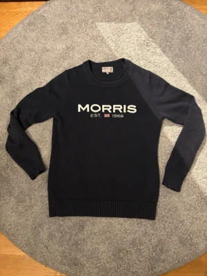 Mörkblå stickad tröja från Morris - Snygg mörkblå stickad tröja från Morris med vit broderad logga och texten 'MORRIS EST. 1968' framtill. Tröjan har rund halsringning och ribbade muddar vid ärmslut och nederkant. Perfekt för en stilren och avslappnad look.